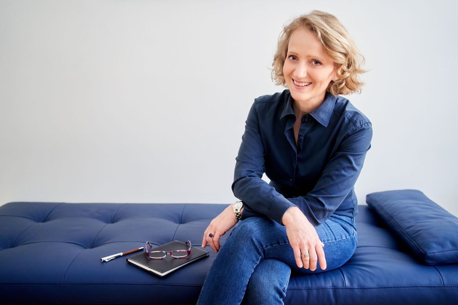 Manuguerra Portrait Fotografie - Eine lächelnde blonde Frau in einem blauen Jeans-Outfit sitzt entspannt auf einer marineblauen Couch.