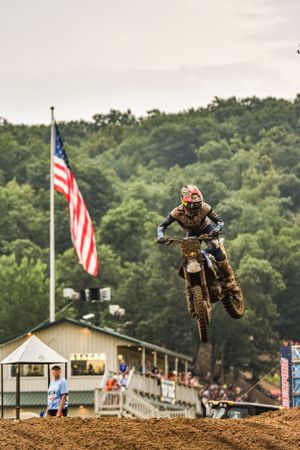 Motocross rider soars through air at race event with American flag and spectator stands visible.
