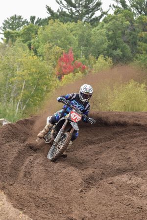 Dirt bike rider navigates a challenging bermed turn on a motocross track surrounded by trees.