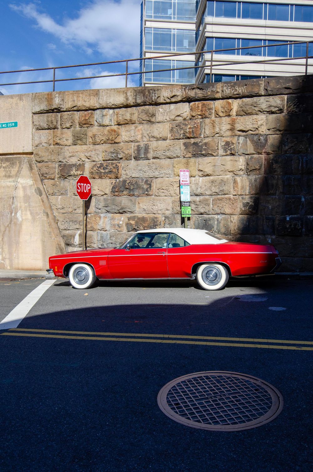 A bright red classic car with white wall tires is parked alongside a stone retaining wall on a city street.