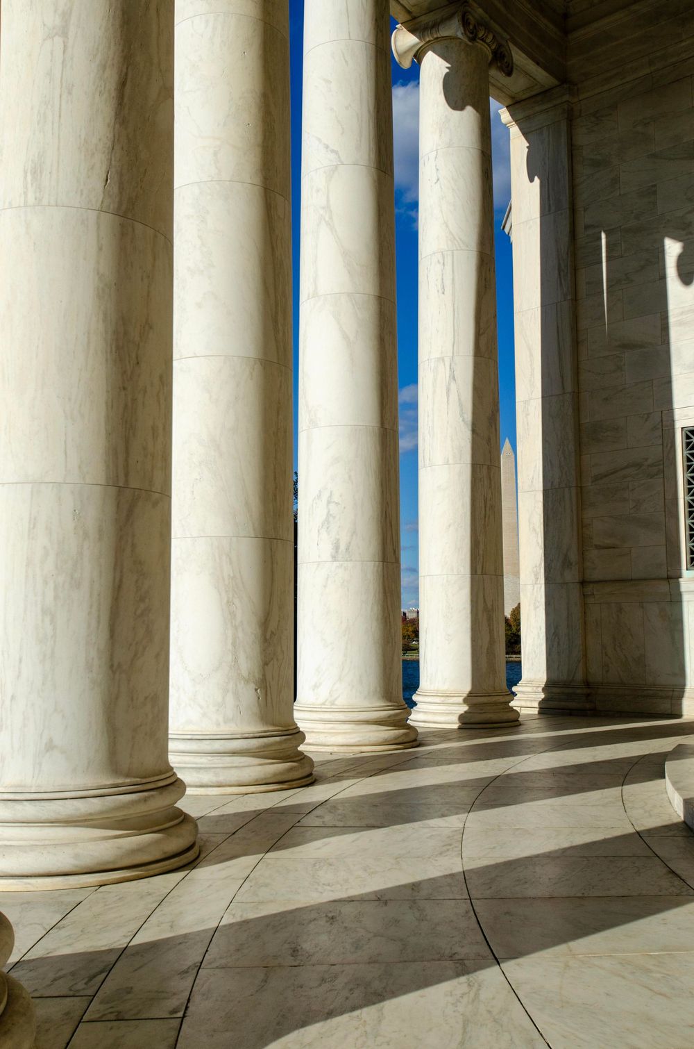 Tall white marble columns cast dramatic shadows on the stone floor of a neoclassical building against a blue sky.