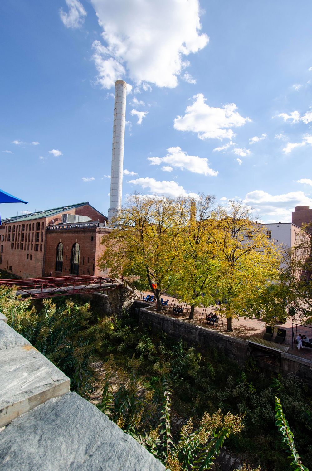 Industrial buildings with brick facades stand against a blue sky with white clouds and autumn trees in the foreground.