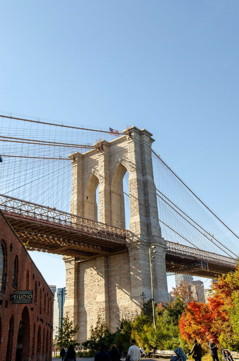 Brooklyn Bridge tower against blue sky with fall foliage in foreground.