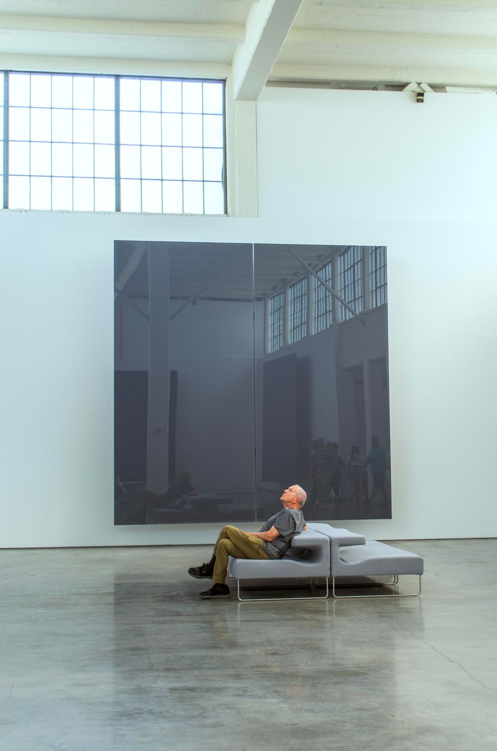 A museum visitor rests on a bench while viewing a large dark minimalist artwork in a bright gallery space.