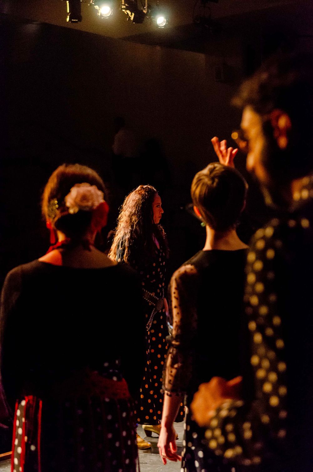 Flamenco dancers in black polka dot dresses perform on a dimly lit stage with dramatic warm lighting.