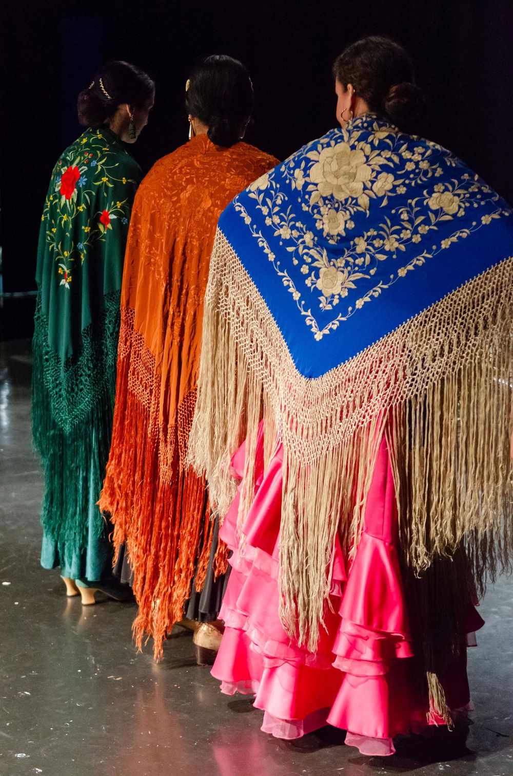 Three flamenco dancers in vibrant ruffled skirts and embroidered fringed shawls stand with their backs to the camera.