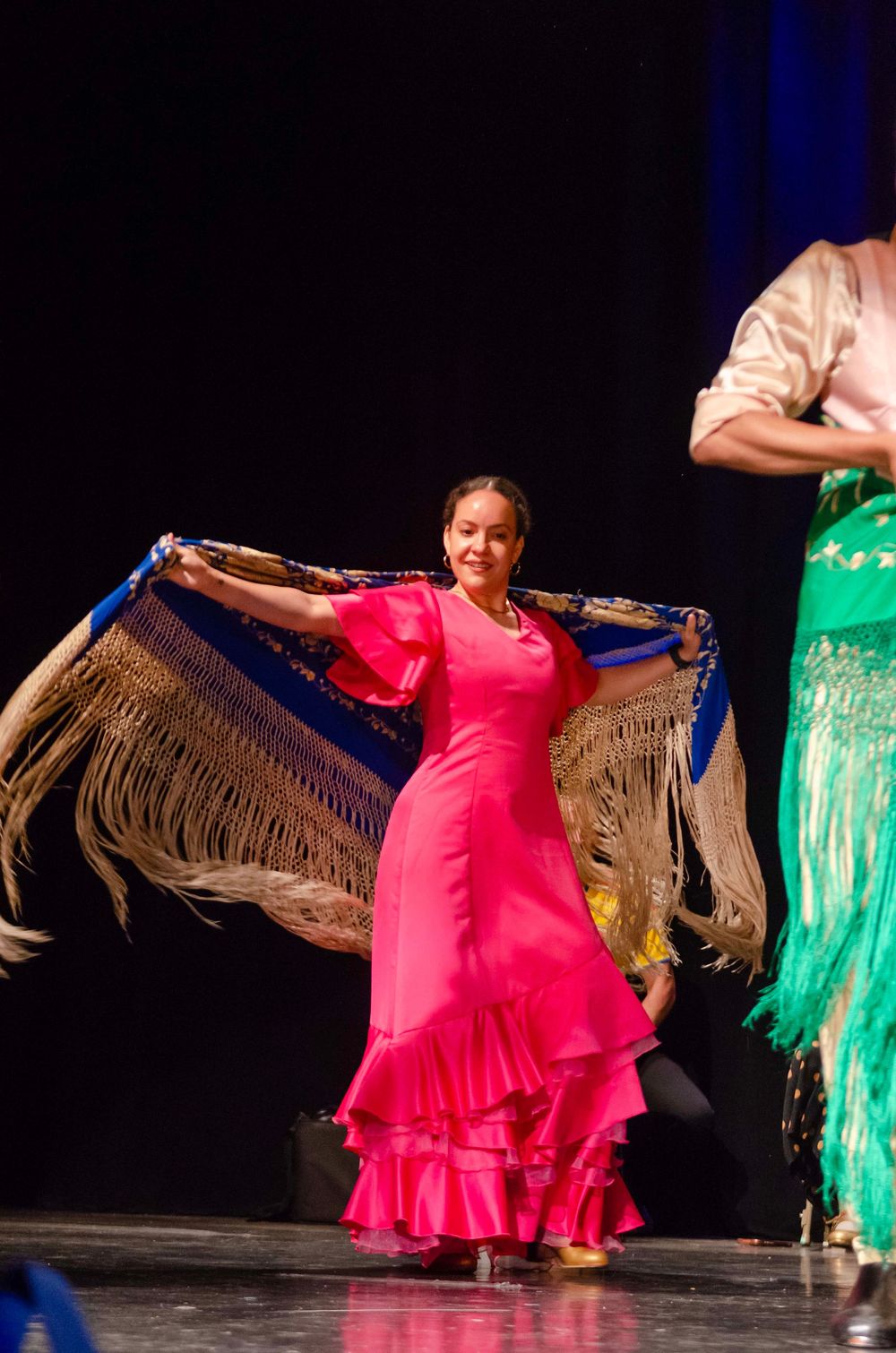 A woman in a pink flamenco dress dances on stage, gracefully holding a fringed shawl with arms outstretched.