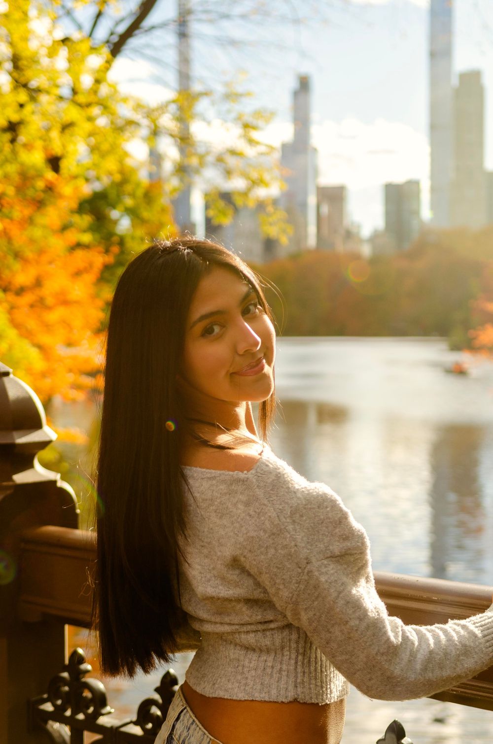 Person in white sweater poses along waterfront railing during autumn sunset with city skyline in background.