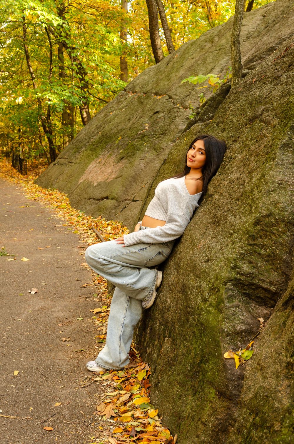 A person in a light sweater leans against a large boulder on an autumn forest path covered in yellow leaves.