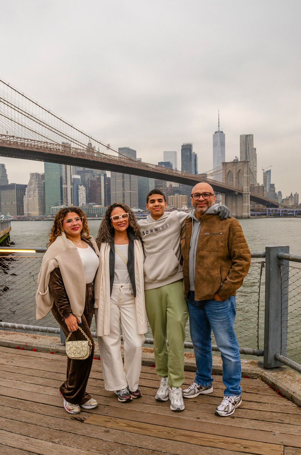 Group of friends posing together on a waterfront boardwalk with Brooklyn Bridge and NYC skyline in background.