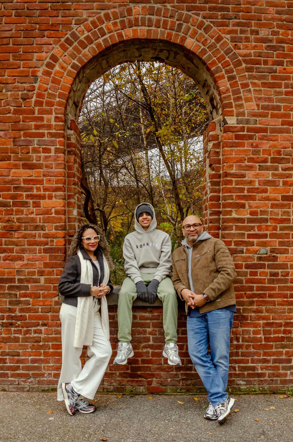 People sit and lean against a red brick wall with arched openings in an autumn outdoor setting.