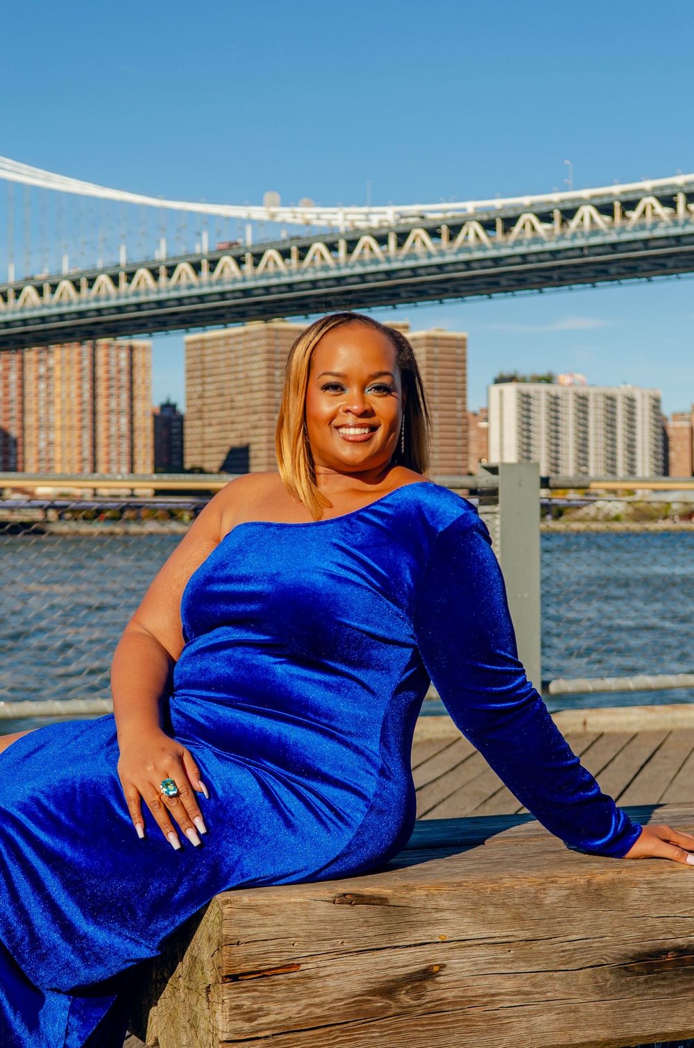 Woman in royal blue velvet dress poses on wooden deck with Manhattan Bridge and East River waterfront in background.