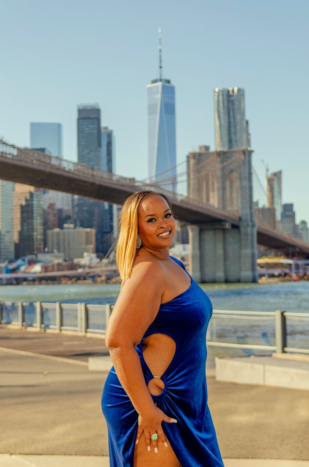 A person in a bright blue dress poses against the New York City skyline and Brooklyn Bridge on a sunny day.