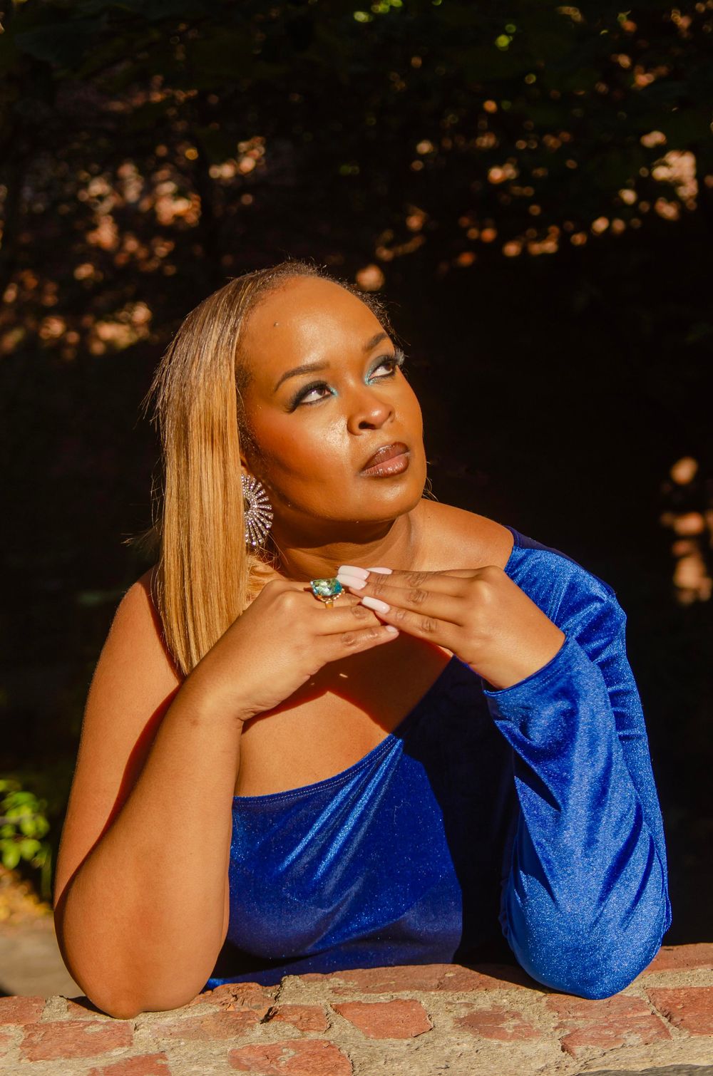 A person in a royal blue velvet dress poses elegantly on a stone surface during golden hour.