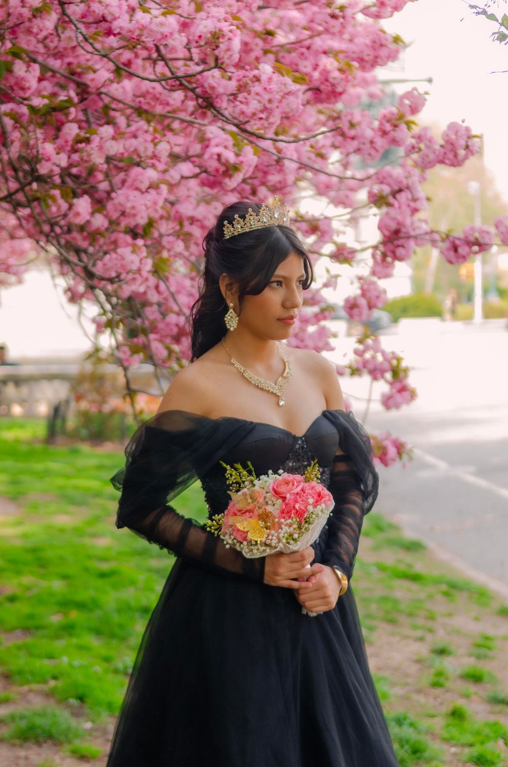 Woman in black off-shoulder gown with tiara holds bouquet under pink cherry blossoms in spring.