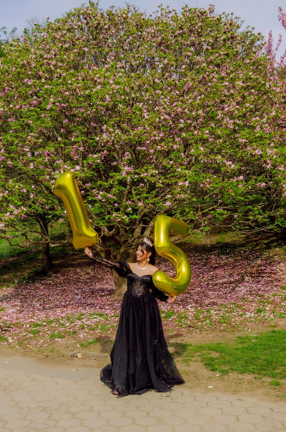 Woman in black gown holding gold number 15 balloons outdoors near blooming pink flowering tree.
