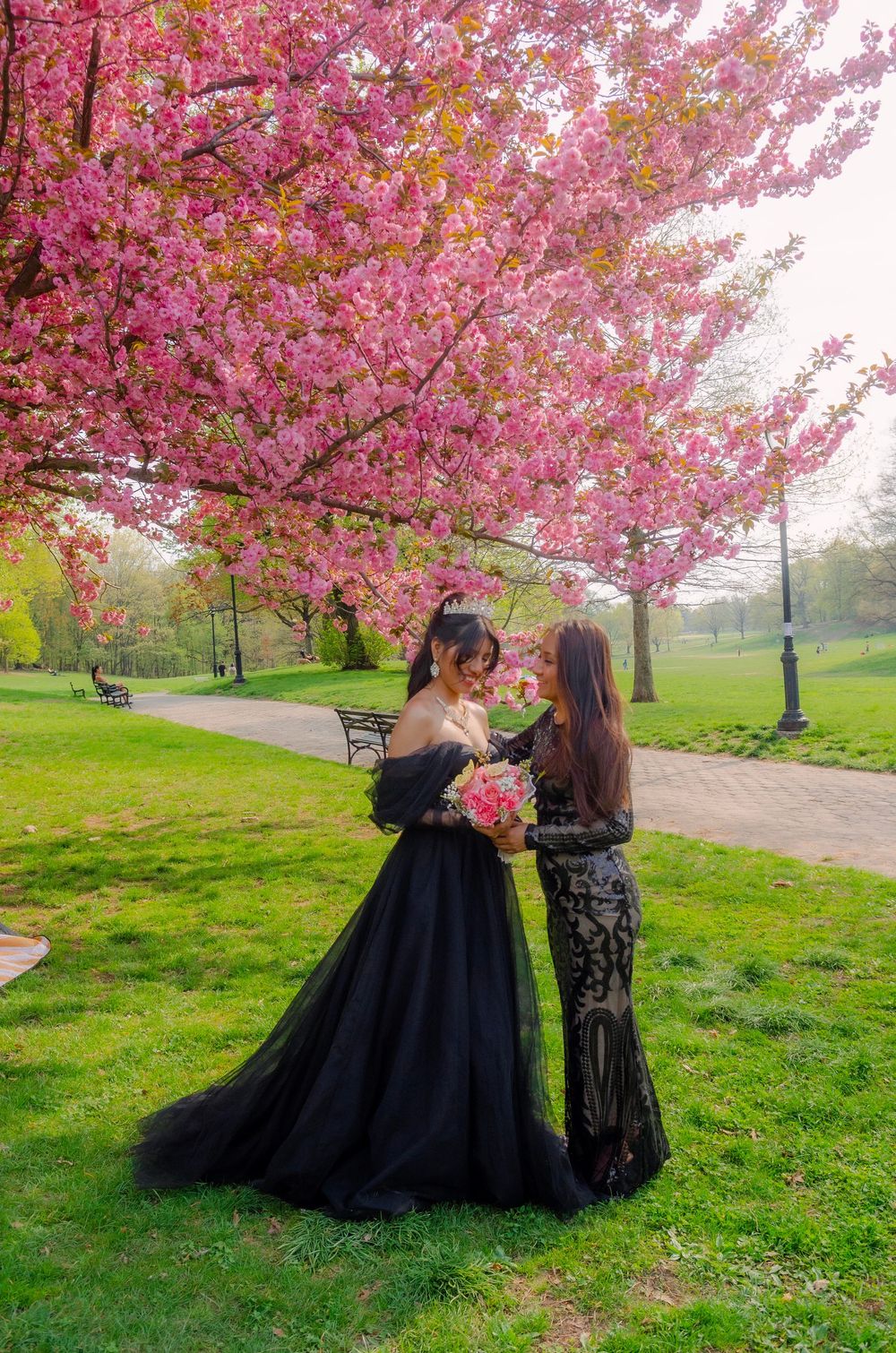 Two women in elegant dresses pose under blooming pink cherry blossom trees in a lush green park.