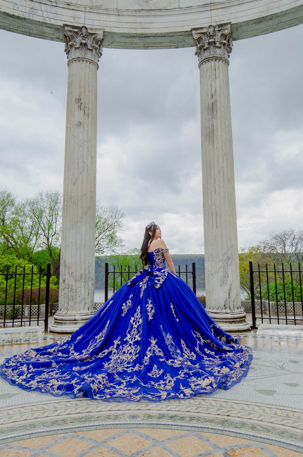 A stunning royal blue quinceañera dress with silver embroidery photographed between classical columns in an outdoor setting.