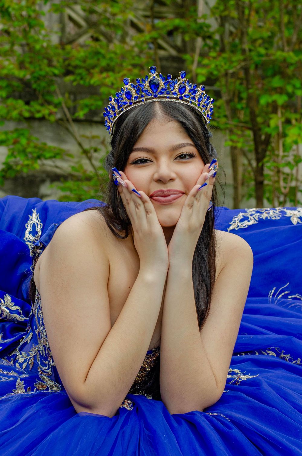 A person in a royal blue quinceañera ball gown with silver embellishments and matching tiara poses in a garden setting.