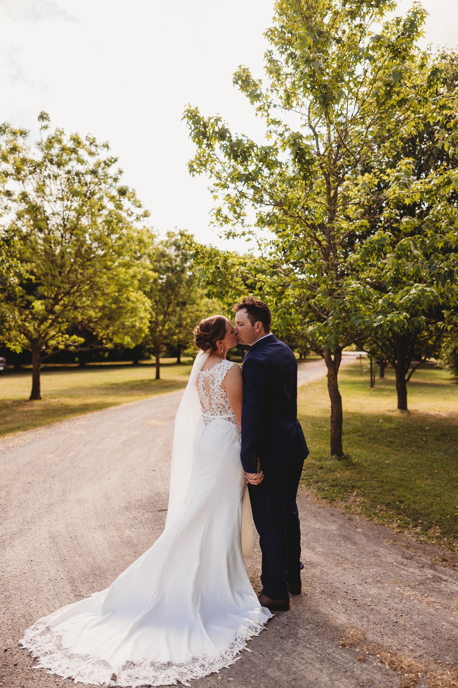 Romantic wedding couple embraces on tree-lined path during golden hour.