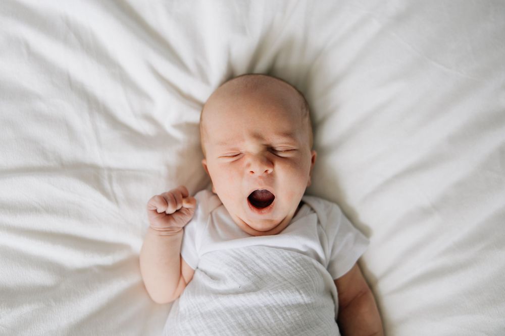 Sleeping newborn baby in white onesie yawning on white bedding.