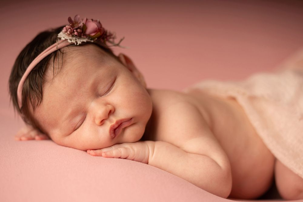 A sleeping newborn baby lies peacefully on soft beige bedding in warm lighting.