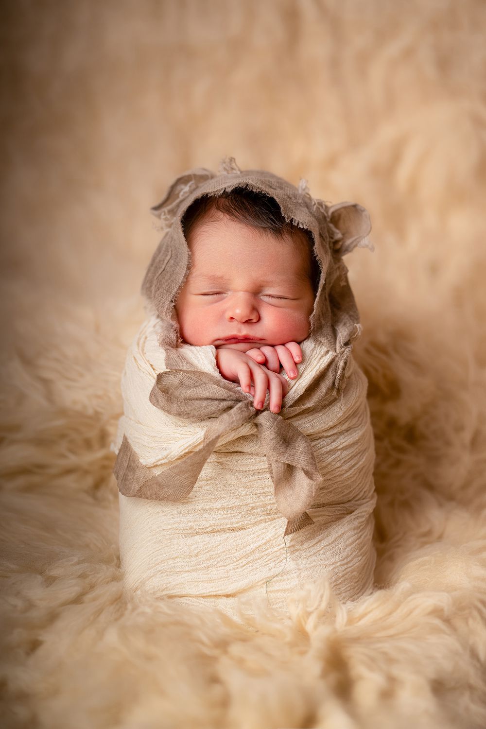 A sleeping newborn baby wrapped in a cream colored swaddle blanket rests peacefully on a soft beige backdrop.