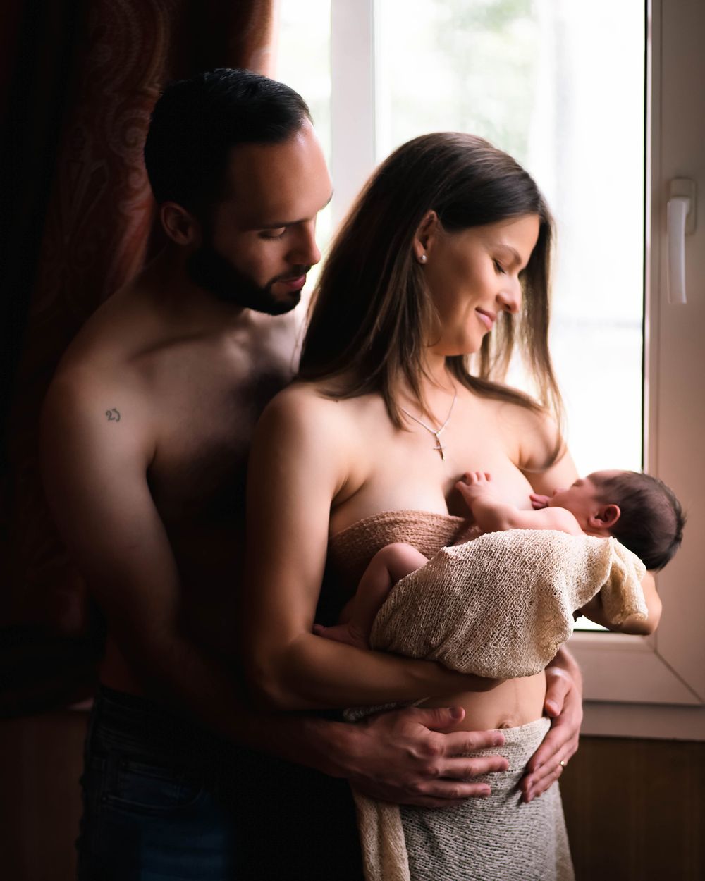 A couple embraces near a window while holding their newborn baby wrapped in a soft blanket.