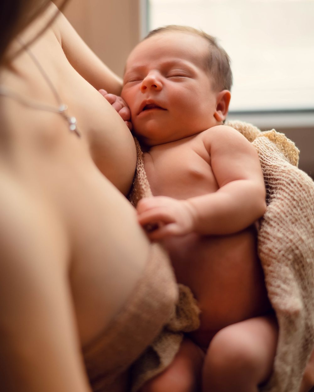 Peaceful sleeping newborn wrapped in beige blanket resting against parent's chest in soft natural lighting.