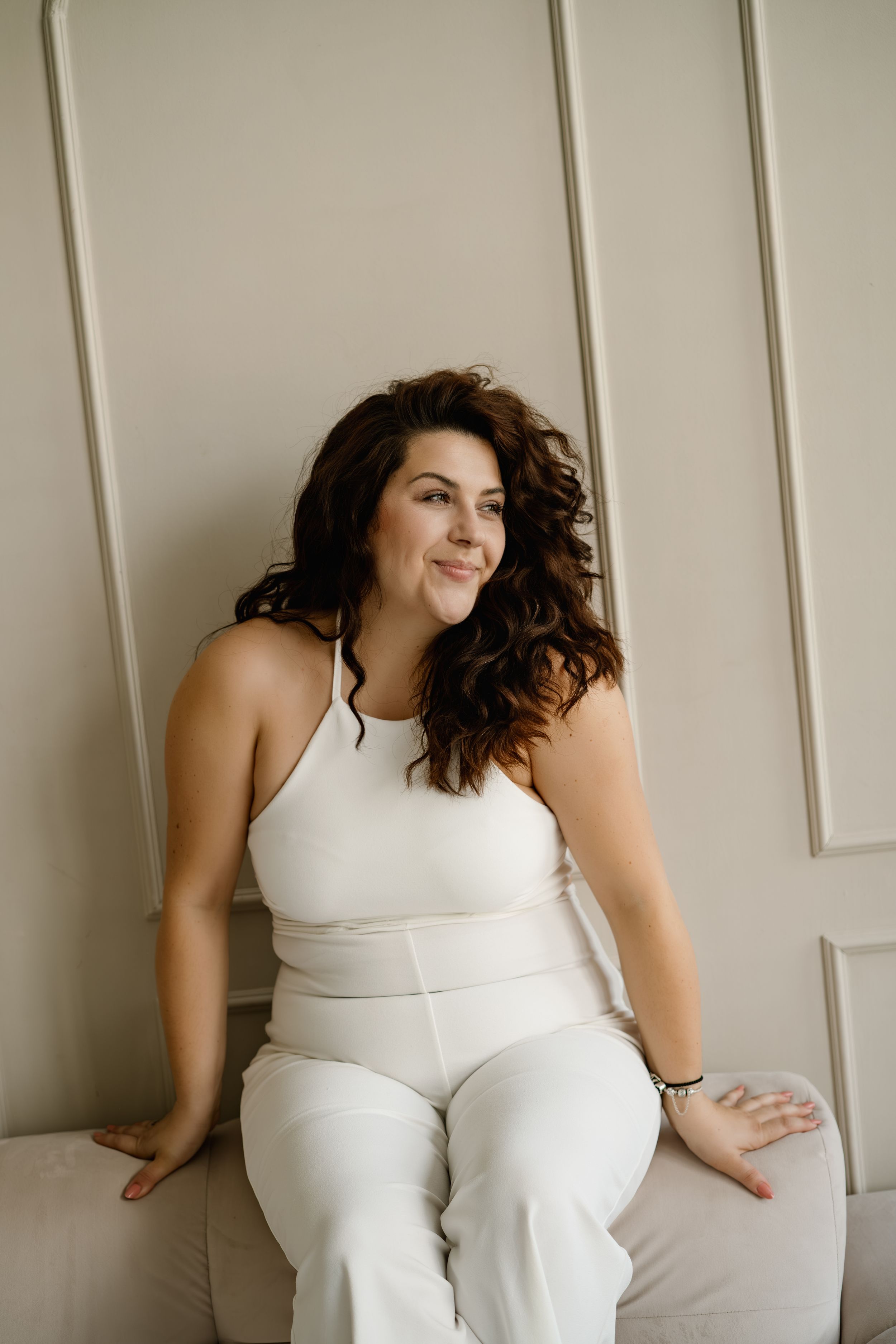 Woman in white eyelet dress sitting against white background with relaxed pose and natural curly hair.