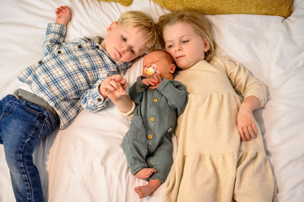 Siblings resting together on a bed with sleeping newborn. Lifestyle newborn photoshoot in Las Vegas. Newborn photographer Las Vegas.