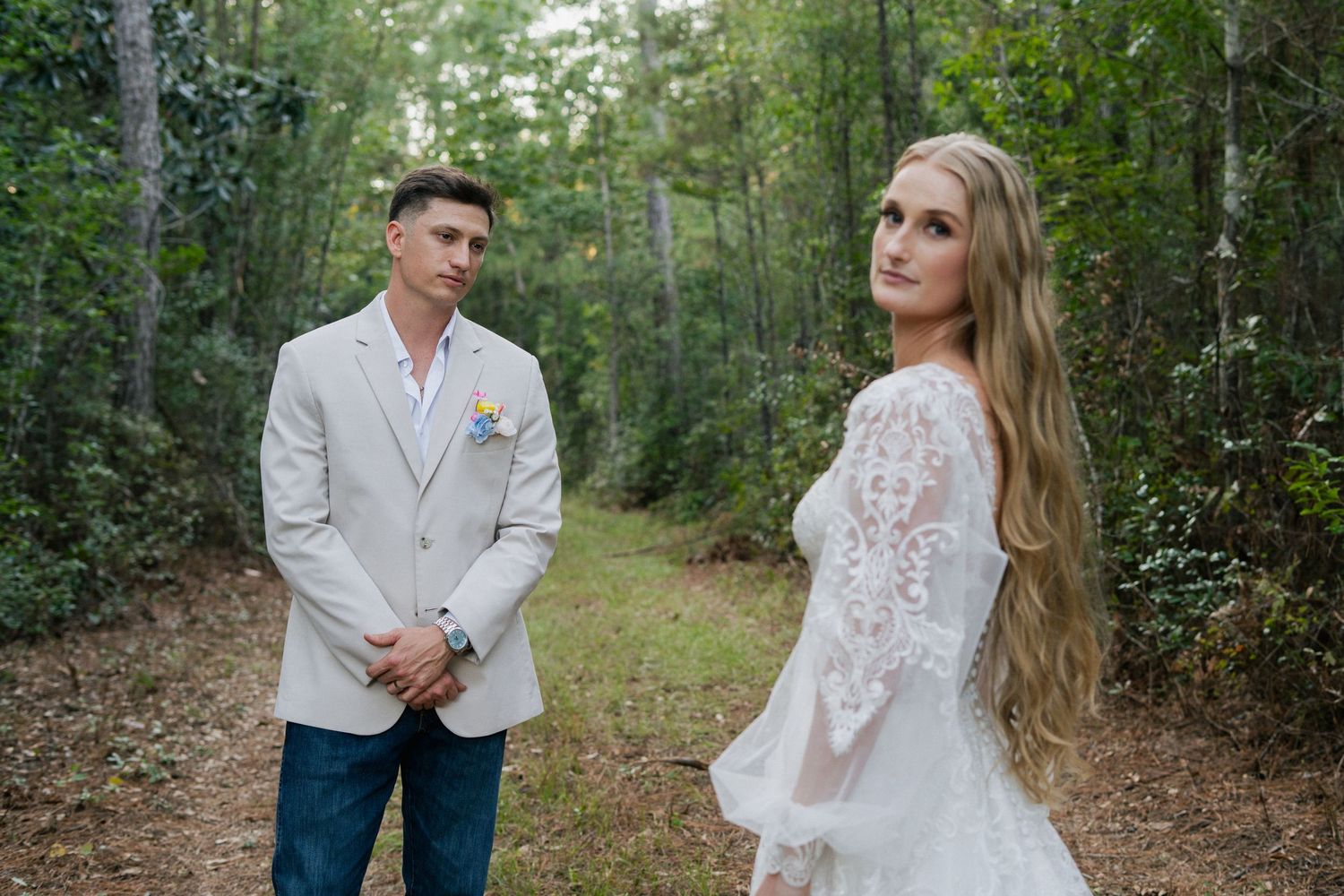 Bride in a long lace wedding dress and groom in a light blazer with jeans pose together on a wooded forest path.