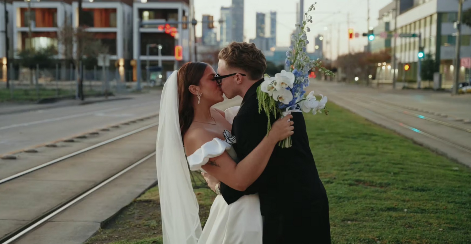 A romantic couple shares a kiss on a city street with tram tracks and urban buildings in the background.