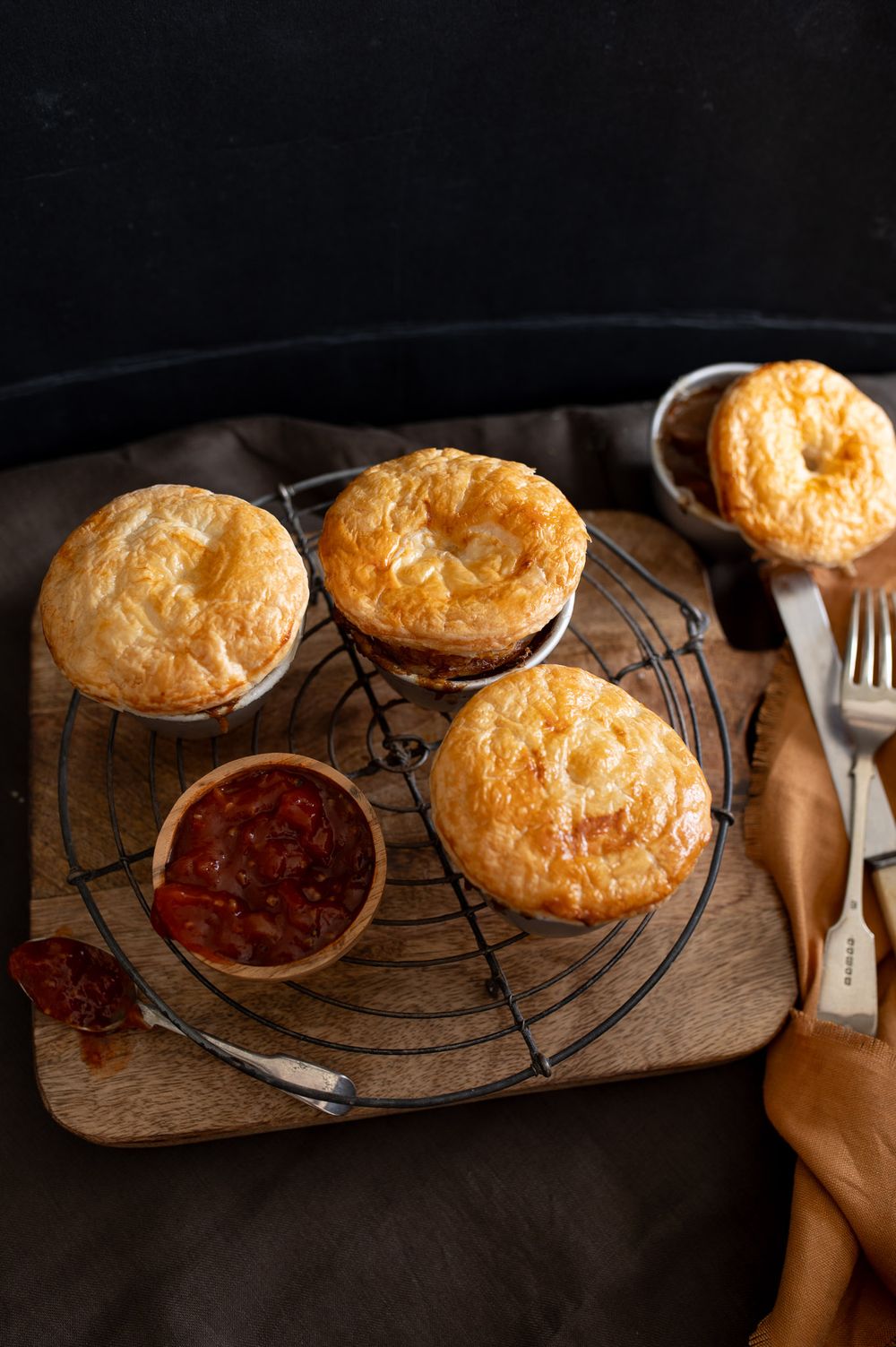 Homemade golden puff pastry meat pies on a cooling rack with red sauce in a wooden bowl on dark background.