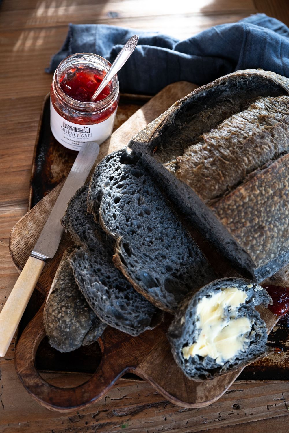 Sliced black charcoal bread with butter and red jam on a rustic wooden board with a spreading knife.