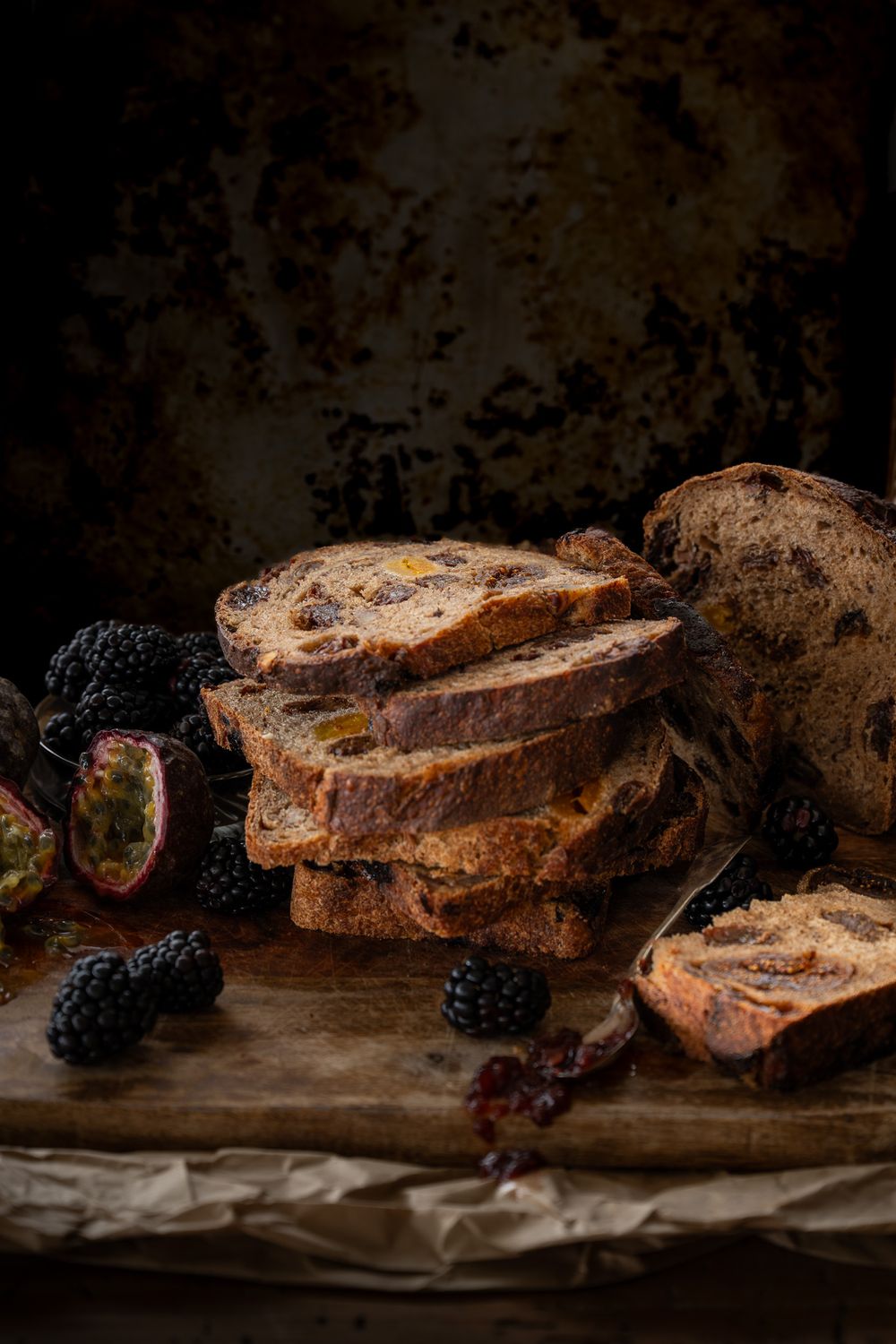 Rustic sliced raisin bread from Barcino's Bread with fresh blackberries arranged on a wooden surface against a dark moody background.