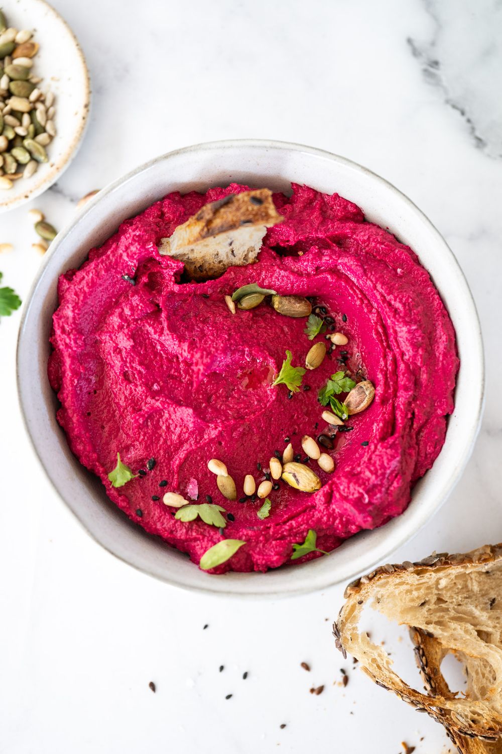 Vibrant pink beet hummus garnished with pumpkin seeds and herbs in a white bowl beside toasted bread slices.