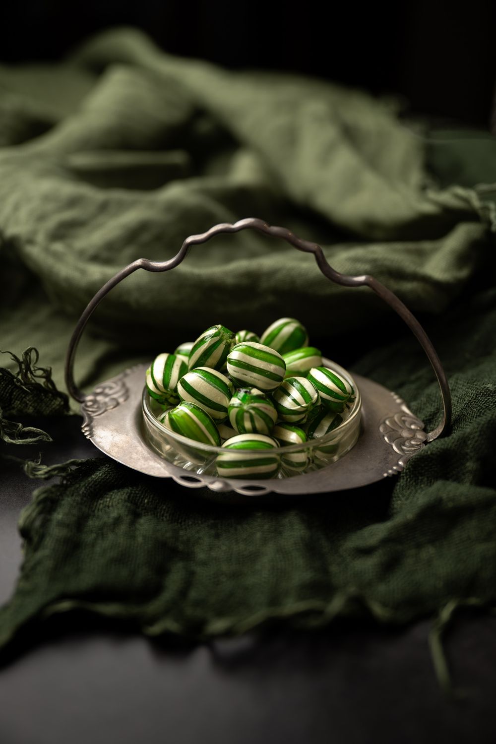 Striped green and white candies arranged on a vintage silver tray with handle against dark fabric background.
