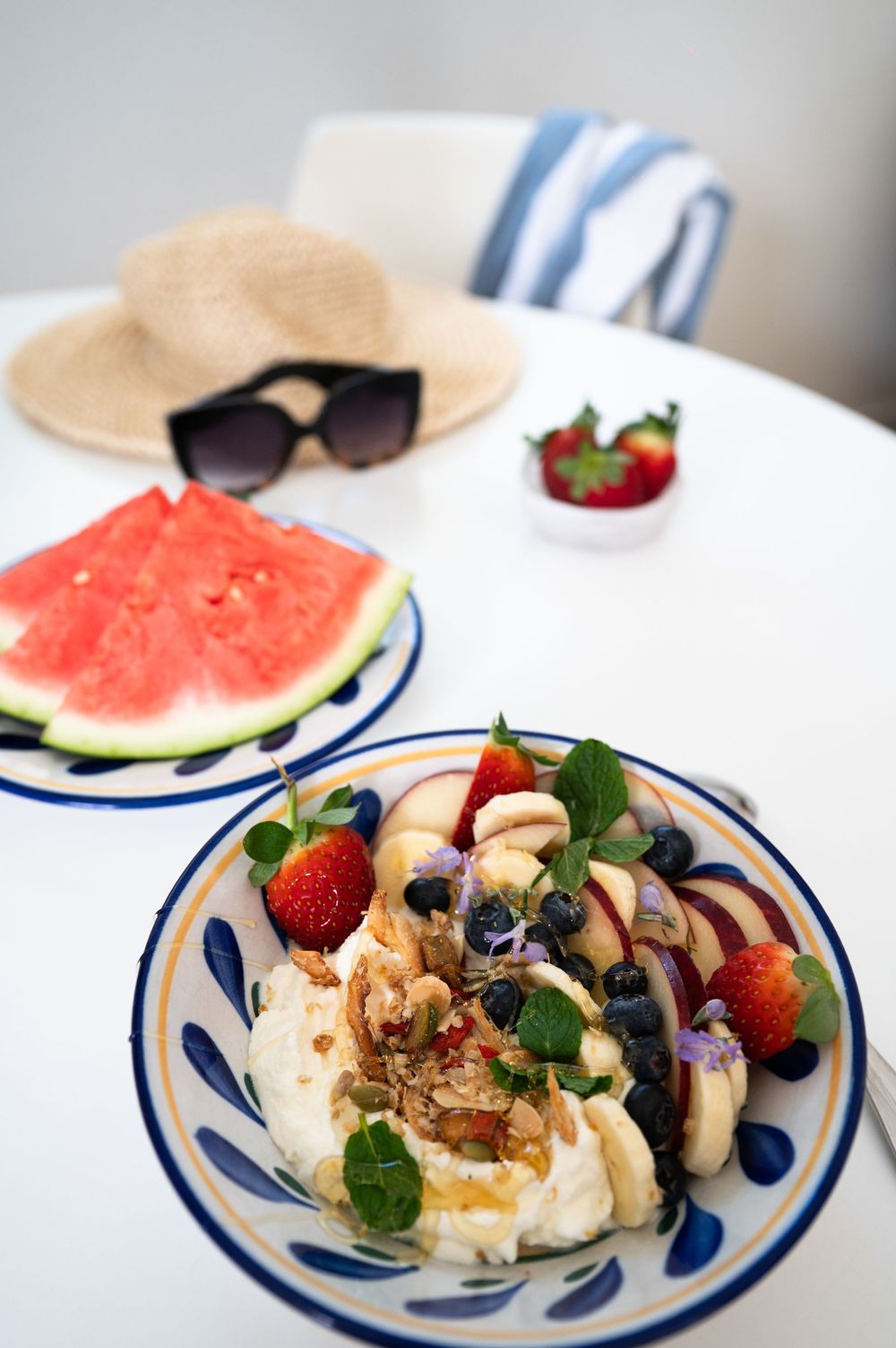 A summer plate with fresh fruit yogurt bowl, watermelon slice, berries and a straw hat with sunglasses in background.