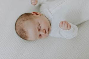 A sleeping baby in white clothes rests peacefully on white bedding.