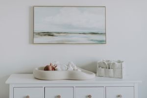 A serene coastal landscape painting hangs above a white dresser decorated with minimalist decor pieces.