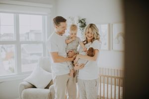 A family in white clothing stands together in a bright, sunlit nursery room near a window.