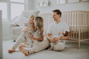 A warm family moment sitting together on the floor of a light-filled nursery with a wooden crib nearby.