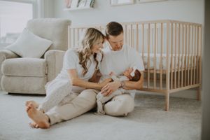 A tender moment between parents and their newborn baby sitting on the floor beside a wooden crib in a bright nursery.
