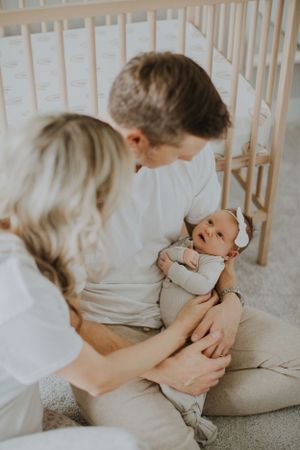 A baby in soft gray laying on a floor by a wooden crib while being cared for with gentle attention.