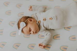 A sleeping baby wearing a white outfit lays on a rainbow patterned sheet.