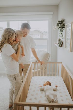 Family in white clothing gathering around wooden crib with baby in a bright, minimalist nursery room.