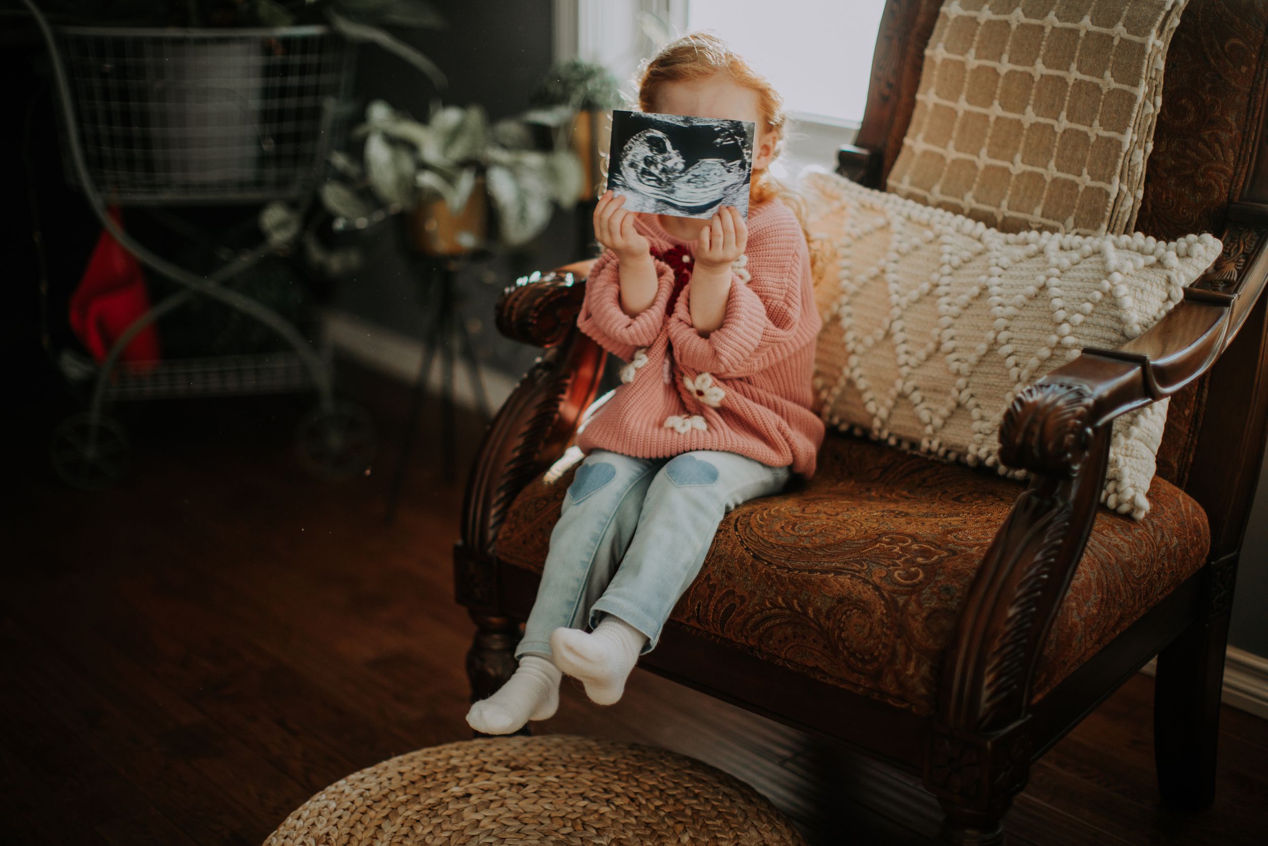 A child in pink sweater and sunglasses sits on vintage brown chair with crocheted cushions on hardwood floor.