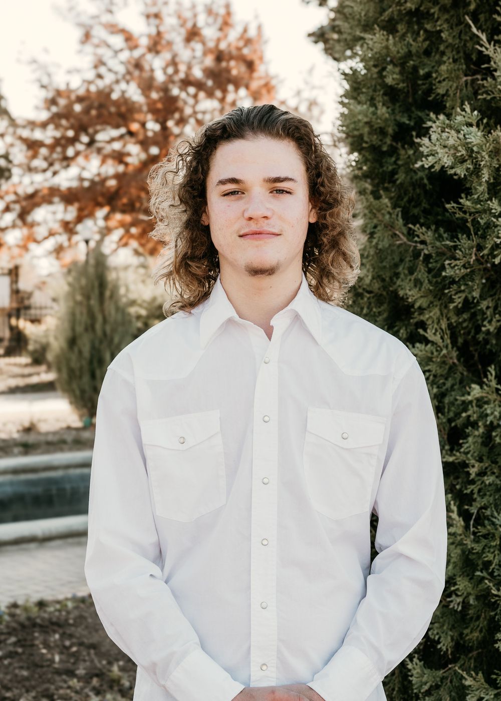 A person in a white button-up shirt poses outdoors during autumn with fall foliage in the background.