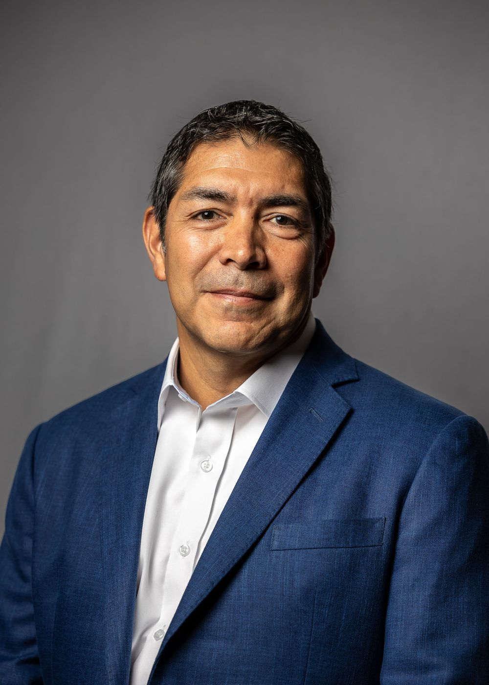 Professional headshot of a person in a navy blue blazer and white dress shirt against a gray background.
